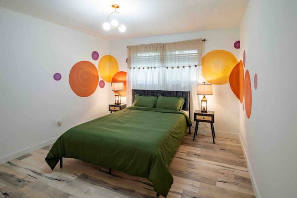 Un dormitorio con una cama verde, dos mesitas de noche con lámparas, una ventana con cortinas y una obra de arte abstracta en la pared.