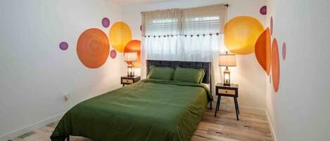 Un dormitorio con una cama verde, dos mesitas de noche con lámparas, una ventana con cortinas y una obra de arte abstracta en la pared.