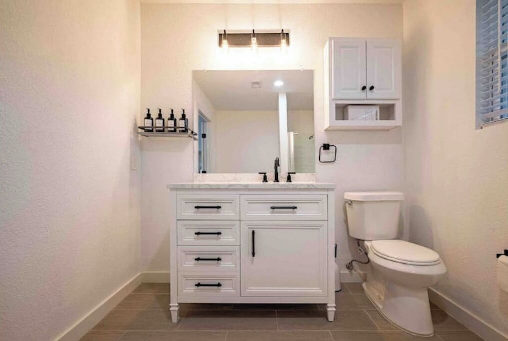 Une salle de bain avec un meuble-lavabo blanc, des toilettes et une étagère remplie de bouteilles.