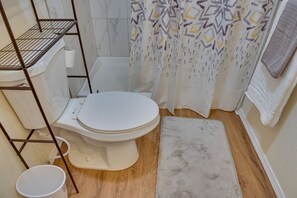 A bathroom with a white toilet, a wooden floor, a white rug, a metal shelf, and a patterned shower curtain. - Soldotna Vacation Rental ~ 8 Mi to Kenai River! (Soldotna)