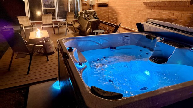 An outdoor hot tub on a wooden deck with chairs and a table, surrounded by a brick wall and string lights. - Spectacular Mountain Views - Hot Tub & Fire Pit (Centennial)