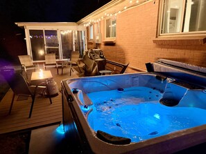 Un bain à remous extérieur installé sur une terrasse en bois, agrémentée de chaises et d’une table, le tout entouré d’un mur de briques et illuminé par des guirlandes lumineuses.