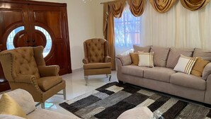 Un salon avec un canapé marron, deux fauteuils, un tapis à motifs et une porte en bois dotée d’une fenêtre ronde.