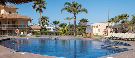 A swimming pool with a lifeguard chair, lounge chairs, palm trees, and a building with a tiled roof.