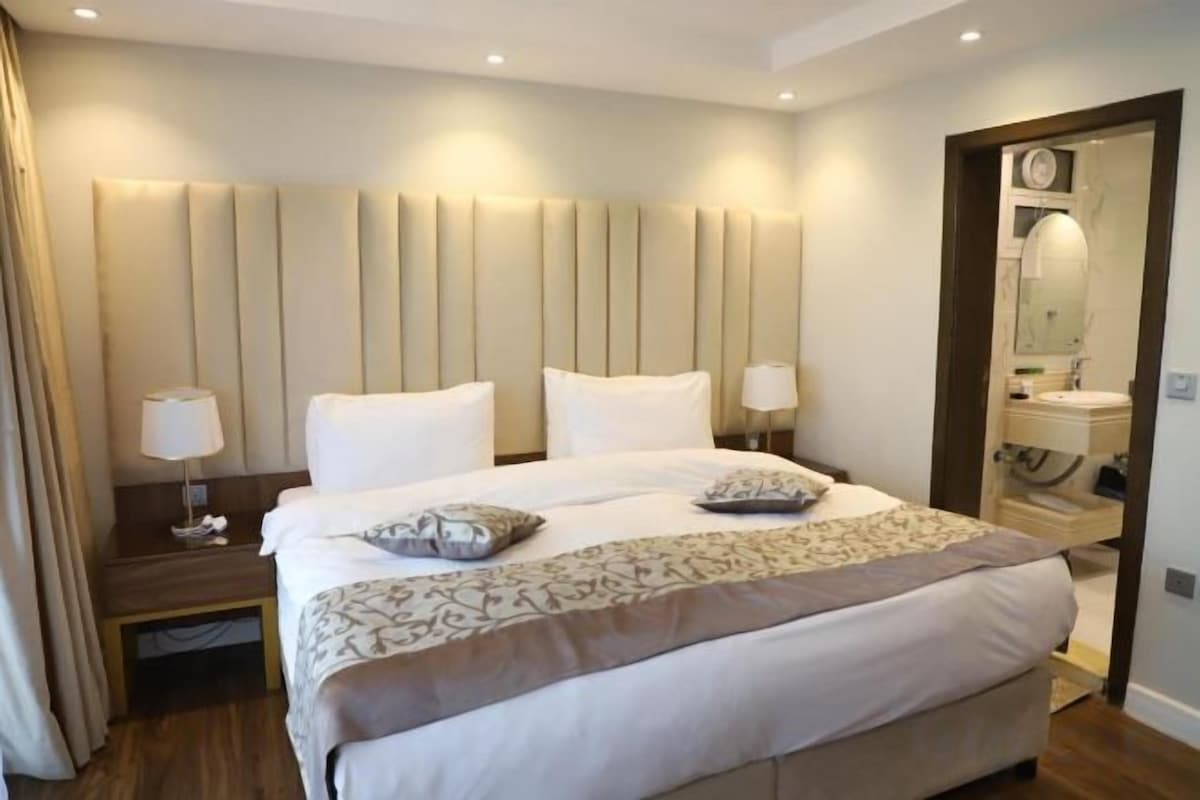 Camera d'albergo con un letto grande, lampade sui comodini, un tavolino da notte e un bagno visibile attraverso una porta aperta.
