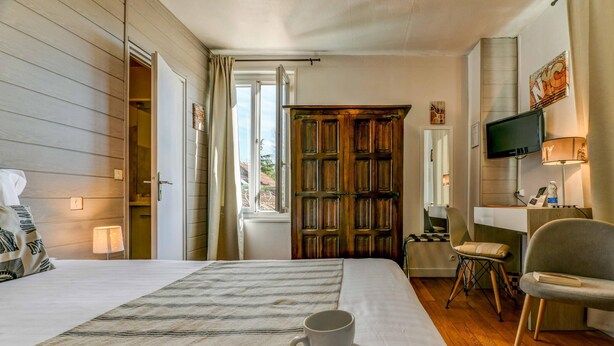 A hotel room with a bed, a desk with a lamp, a television, and a window with curtains. - Logis Hostellerie du Neez (Gan)