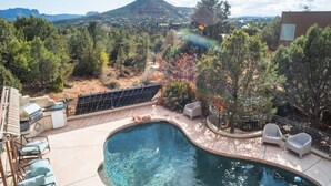 A swimming pool with a curved edge, surrounded by outdoor furniture and a view of a mountainous landscape.