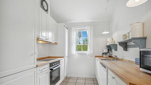 Ett kompakt kök med vita skåp, en träbänkskiva, en inbyggd ugn, ett handfat och ett fönster med gardiner.
