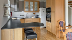 A modern kitchen with wooden cabinets, a black countertop, and a built-in oven. There are two black bar stools and a wooden chair with a striped cushion. - Awesome home in Farsund with WiFi (Farsund)