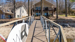 Eine Holzbrücke führt zu einem zweigeschossigen Gebäude mit Balkon, umgeben von Bäumen und einem kleinen Bach.