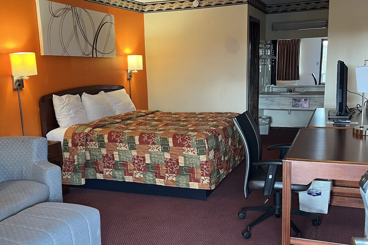 A hotel room with a bed, a desk, a chair, a sofa, and a bathroom visible through an open door.