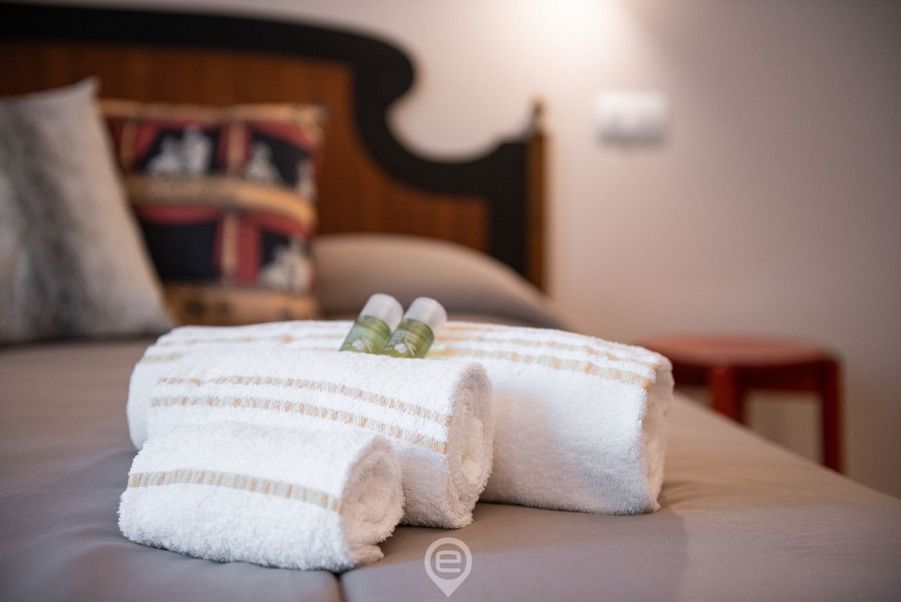 A neatly made bed with a rolled white towel and a striped towel, a small wooden nightstand, and a headboard with a decorative pillow.