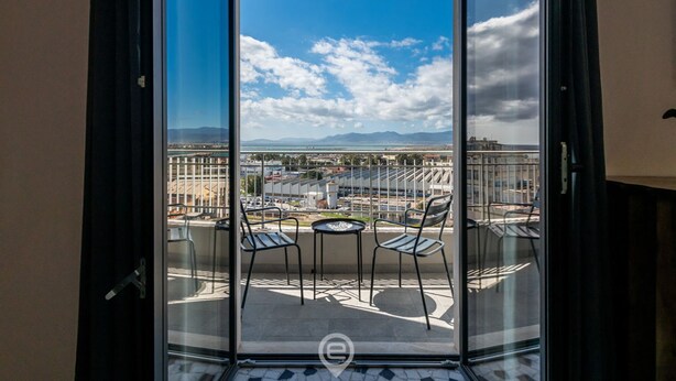 A balcony with a glass door, two chairs, a small table, and a view of a cityscape with mountains in the distance. - Harbor View Apartment (Cagliari)