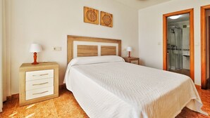 Une chambre à coucher avec un lit, une tête de lit en bois, des tables de chevet, une commode, une lampe, une œuvre d’art accrochée au mur et une porte de salle de bain.