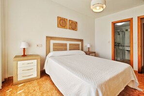 A bedroom with a bed, wooden headboard, bedside tables, a nightstand, a lamp, a wall-mounted artwork, and a bathroom door.