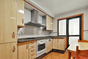 Cuina amb armaris de fusta, forn integrat, finestra amb cortines i una taula de dinar.