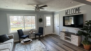Ein geräumiges Wohnzimmer mit einem grauen Sofa, zwei Sesseln, einem Couchtisch, einem großen Fernseher und einem Schild mit der Aufschrift 'WELCOME'.
