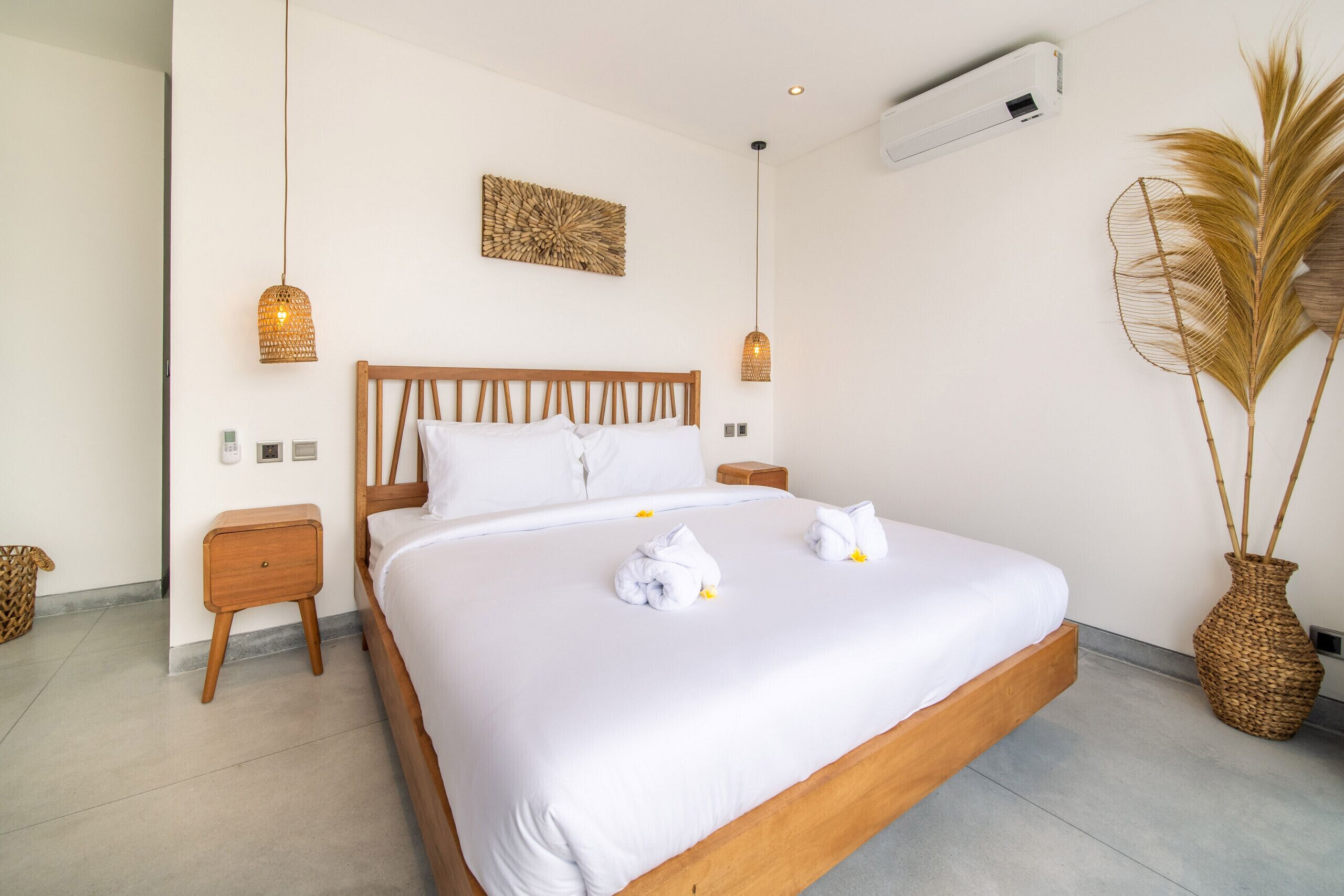 Een keurig opgemaakt bed met witte lakens en twee handdoeken, een houten hoofdbord, nachtkastjes, een wandairconditioner en een decoratieve geweven vaas.