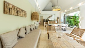 Una sala de estar moderna con sofá, mesa de comedor y sillas. Se observa un área de cocina al fondo y un ventanal con plantas.