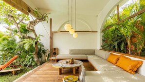 A modern living room with a wooden floor, a round coffee table, a beige sofa, and a small orange chair. There is a large window with a view of greenery outside. - Tropical Garden | Three Bedrooms | Walk to Cafés (Kerobokan)