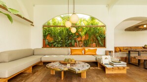 A modern living room with a large arched window, a wooden coffee table, a sofa with orange cushions, and a dining area with a wooden table and chairs. - Tropical Garden | Three Bedrooms | Walk to Cafés (Kerobokan)