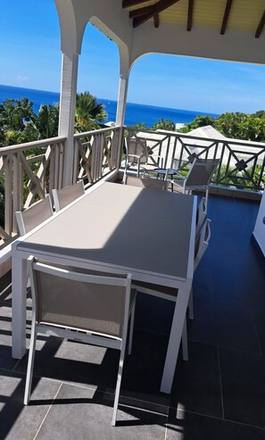 A covered outdoor patio with a table and chairs, overlooking a scenic ocean view.