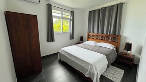 Une chambre à coucher avec une tête de lit en bois, un lit avec une literie blanche, une petite table de chevet, une commode, une fenêtre avec des rideaux et une porte.
