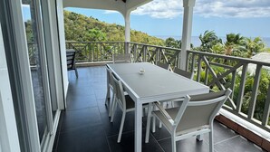Un espace extérieur abrité, avec une table et des chaises blanches, une porte vitrée et une vue sur l’océan et les collines.