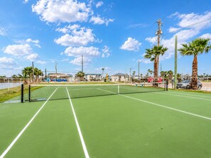 A tennis court with a net, surrounded by a fence, palm trees, and a clear sky. - Canal | Hot Tub | Large Deck | Private Dock | Pets (Galveston)