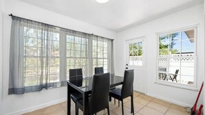 Une pièce avec une table en verre et quatre chaises, de grandes fenêtres avec des rideaux légers, une porte blanche et une vue sur un patio aménagé avec du mobilier d’extérieur.