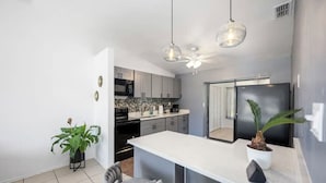 Une cuisine moderne avec un plan de travail blanc, des armoires grises et un réfrigérateur noir. On y trouve deux lampes suspendues, un ventilateur de plafond et une plante en pot sur le plan de travail.