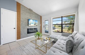 Un salon moderne avec un canapé gris, une table basse en verre agrémentée d’une plante, un téléviseur fixé au mur et une grande fenêtre donnant sur l’extérieur.