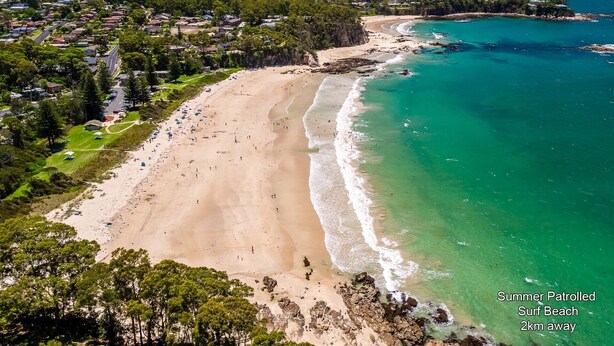 Vue aérienne d’une plage aux eaux turquoise et limpides, bordée d’un rivage sablonneux, avec une zone résidentielle en arrière-plan.