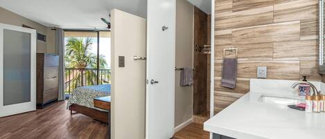 A modern hotel room with a wooden floor, a bed, a bathroom with a wooden wall, and a balcony with a view of palm trees.