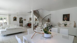 A modern living room with a white sofa, a dining table with orchids, and a spiral staircase. - Peaceful Villa with Infinity Pool, 10' from Center (Antibes)