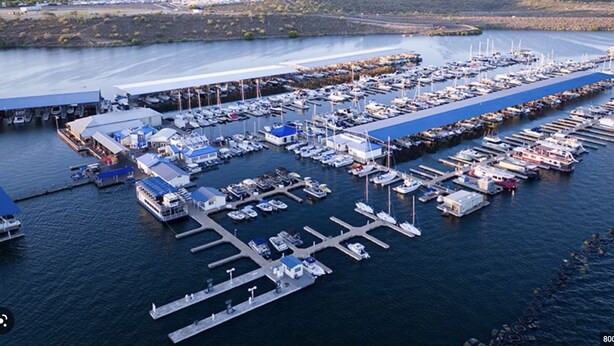 Vista aérea de uma marina com vários barcos atracados, uma grande estrutura coberta e uma cadeia montanhosa ao longe.