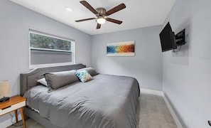 Ein Schlafzimmer mit einem Bett, einem Deckenventilator, einem Fenster, einem an der Wand montierten Fernseher und einem farbenfrohen abstrakten Gemälde.
