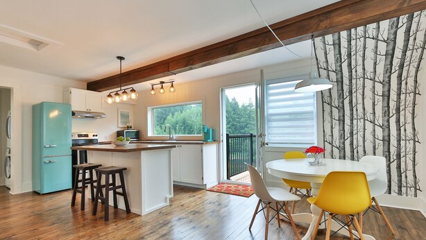 A modern kitchen with a teal refrigerator, wooden flooring, and a dining area with a white table and yellow chairs. - Charming 2 bedroom rental unit in Knowlton village (Knowlton)