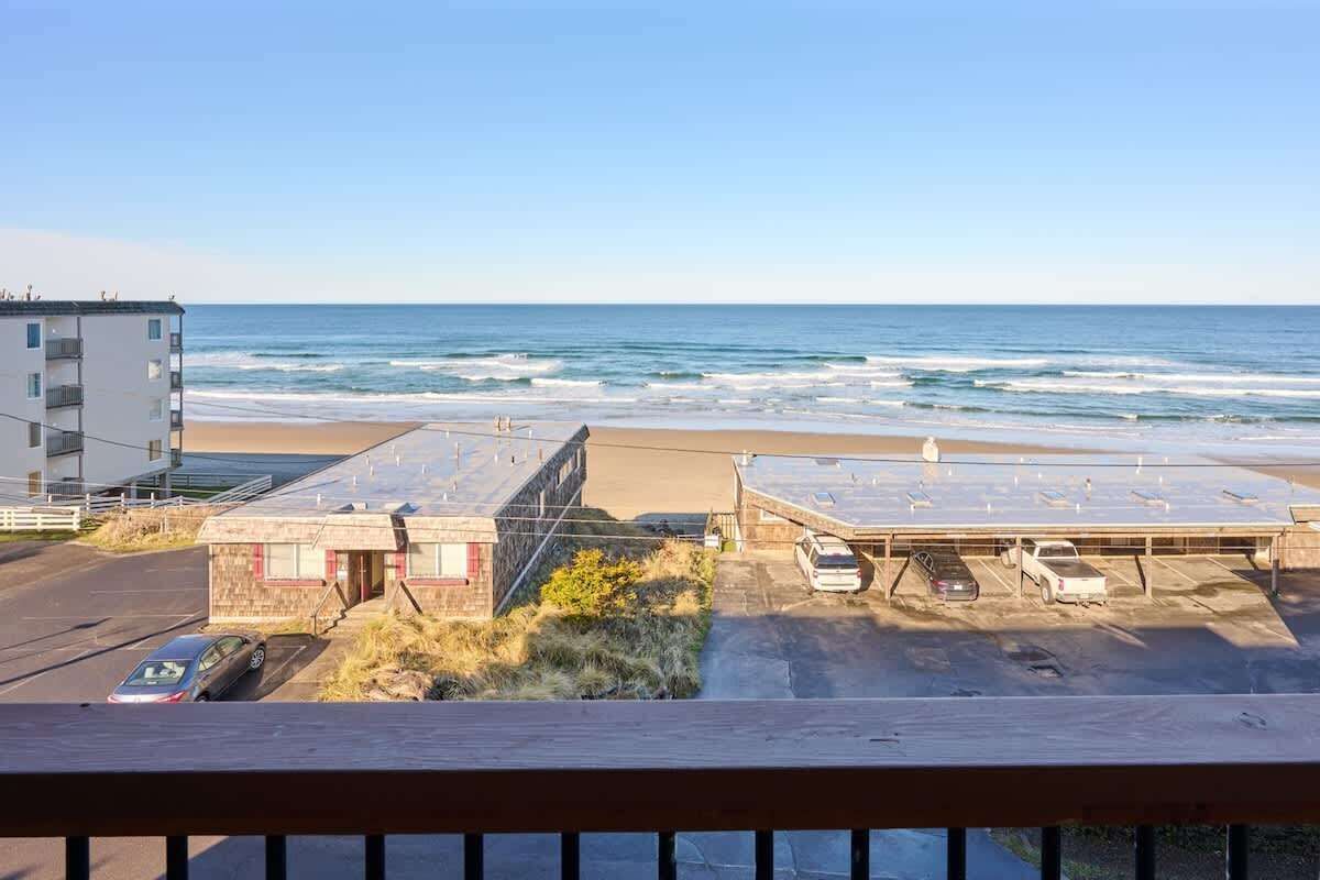 A beach view with a parking lot, a building with a red door, and a multi-story structure in the background.