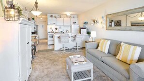 Ein Wohnzimmer mit Sofa, Hocker und Sesseln, im Hintergrund eine Küche und ein großer Spiegel an der Wand.