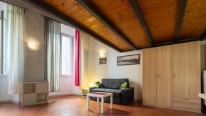 A room with a wooden ceiling, a sofa, a small table, a cabinet, and a window with curtains. - Palazzo Vecchio Place by Mmega (Firenze)
