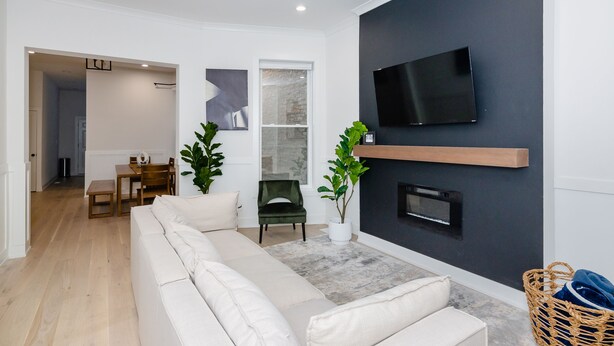 A modern living room with a sofa, a dining table with chairs, a TV mounted on the wall, and a fireplace.