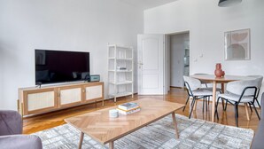 Una sala moderna con un televisor de pantalla plana, una mesa de centro de madera con libros, un estante blanco y un comedor con mesa y sillas.
