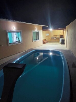 A swimming pool with a built-in hot tub, a small kitchen area with a brick oven, and a seating area with a table and chairs.
