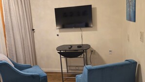 A room with a flat-screen TV mounted on the wall, a small table with a remote control, two blue chairs, and a wooden floor.