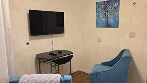 A room with a flat-screen TV mounted on the wall, a small table with a chair, a blue armchair, and a painting of a tree.