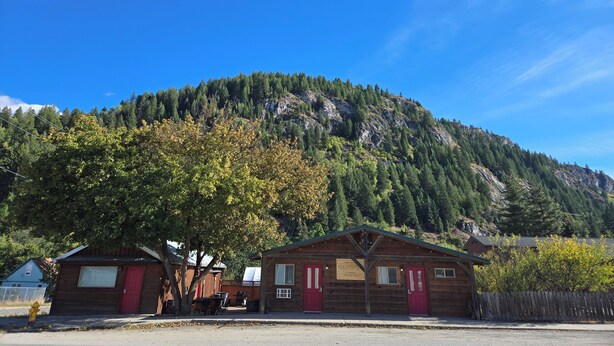 A cabin with a red door and a small porch, surrounded by trees and a mountain backdrop.