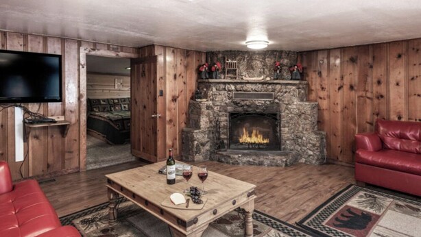 A cozy living room with a stone fireplace, wooden walls, a red leather sofa, and a wooden coffee table with wine glasses and cheese. - Coyote - Upper Canyon w Priv Hot Tub, Big Decks, Wood Fireplace (Ruidoso)