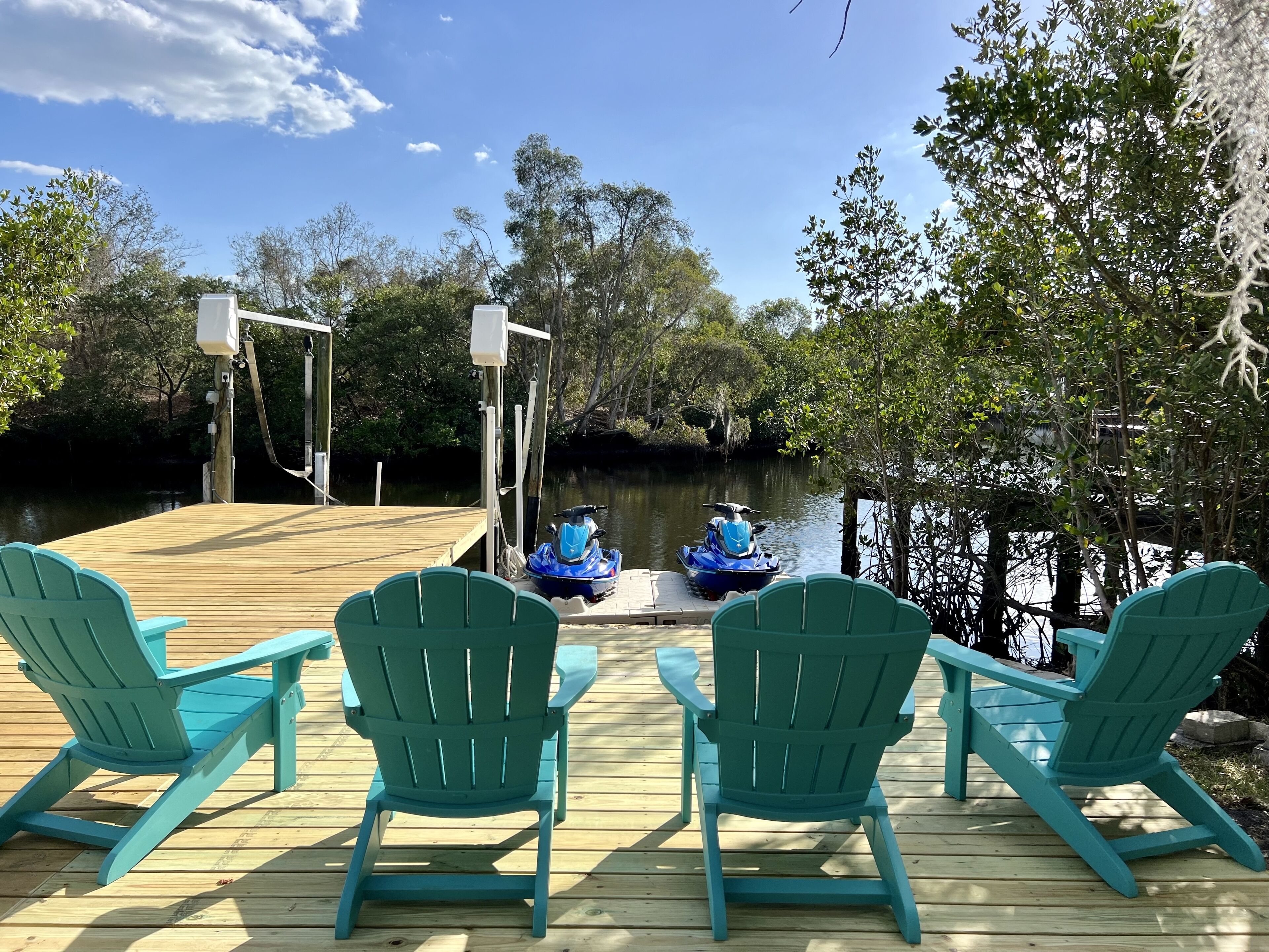 Une terrasse en bois avec des fauteuils Adirondack verts et un jet ski amarré au bord de l’eau.