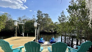Une terrasse en bois avec des fauteuils Adirondack verts et un jet ski amarré au bord de l’eau.